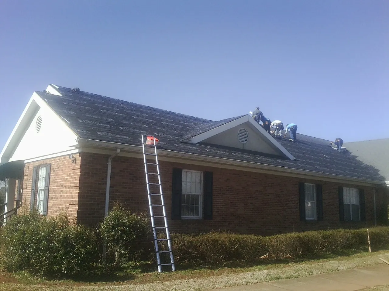 Crew tearing shingles off a brick ranch roof with a ladder set — Emergency Roof Repair work by our crew in Evangeline