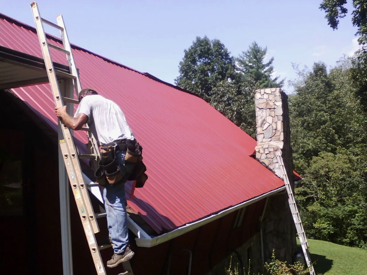Roofer on a ladder beside a red metal roof and stone chimney — Soffit & Fascia Repair by Roofing Brothers in Evangeline